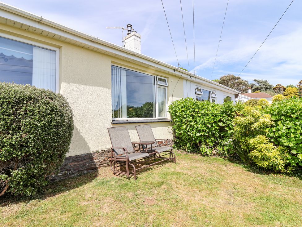 A garden with two chairs and a small table in front of a house with bushes and windows at Ambermist in Churchstow