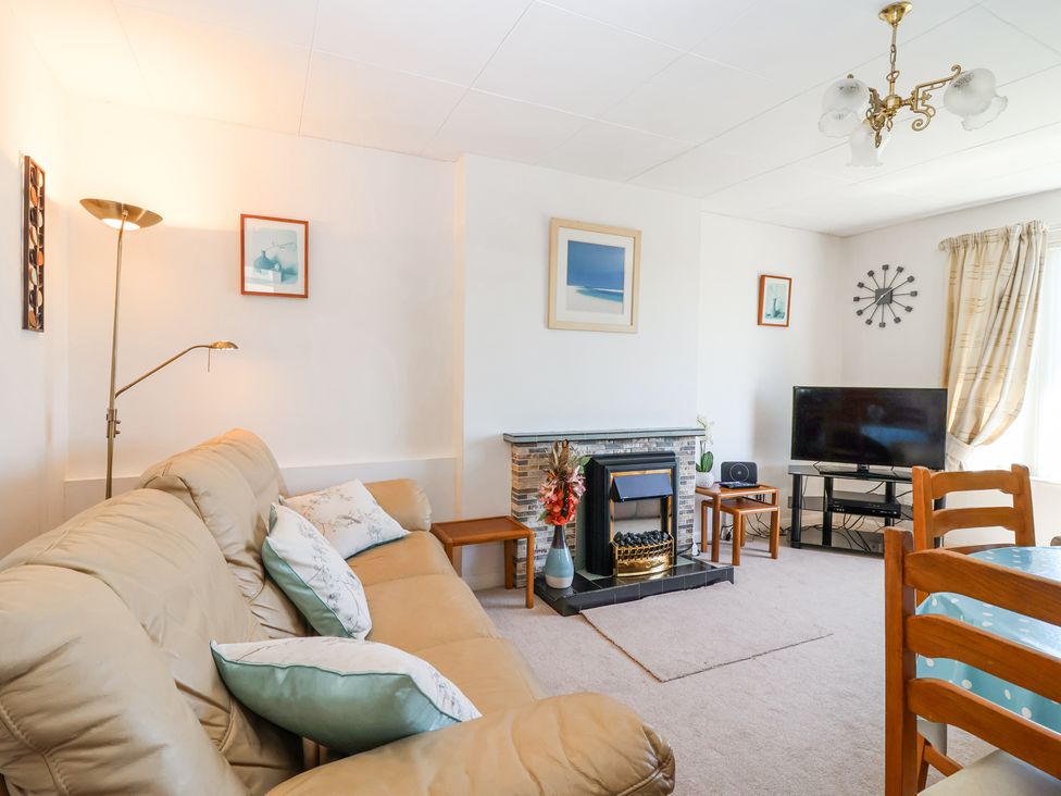 A living room with a beige sofa, floor lamp, fireplace, television on a stand, wall clock, and wooden dining chairs at Ambermist in Churchstow