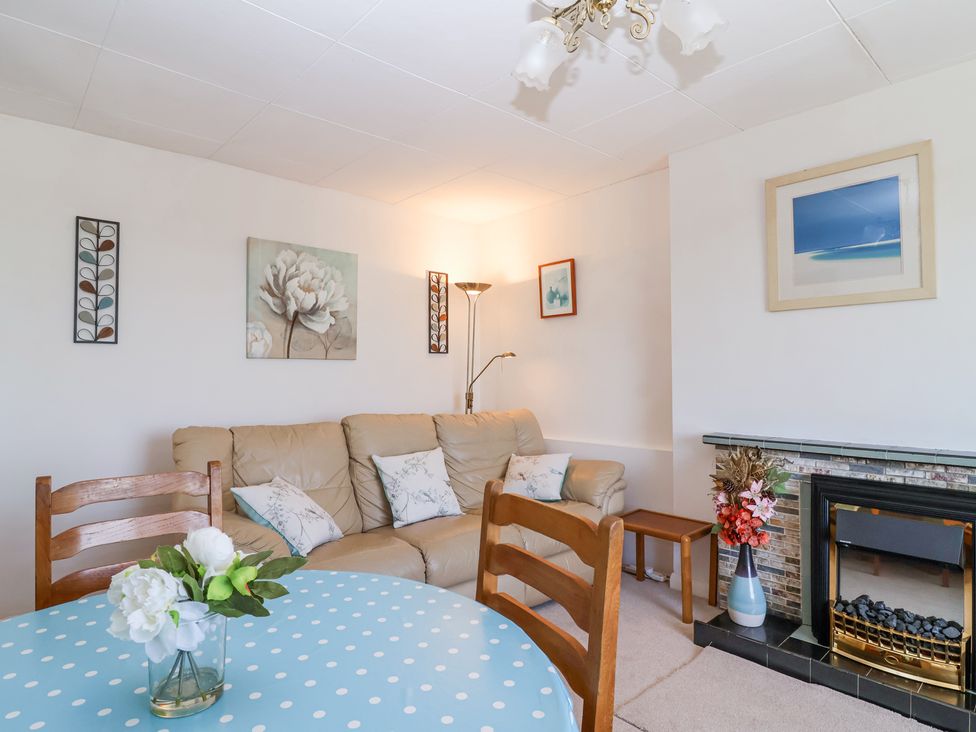 A living room with a beige sofa and floral cushions a round table with a blue polka dot tablecloth a fireplace with a vase of flowers and framed pictures on white walls at Ambermist in Churchstow