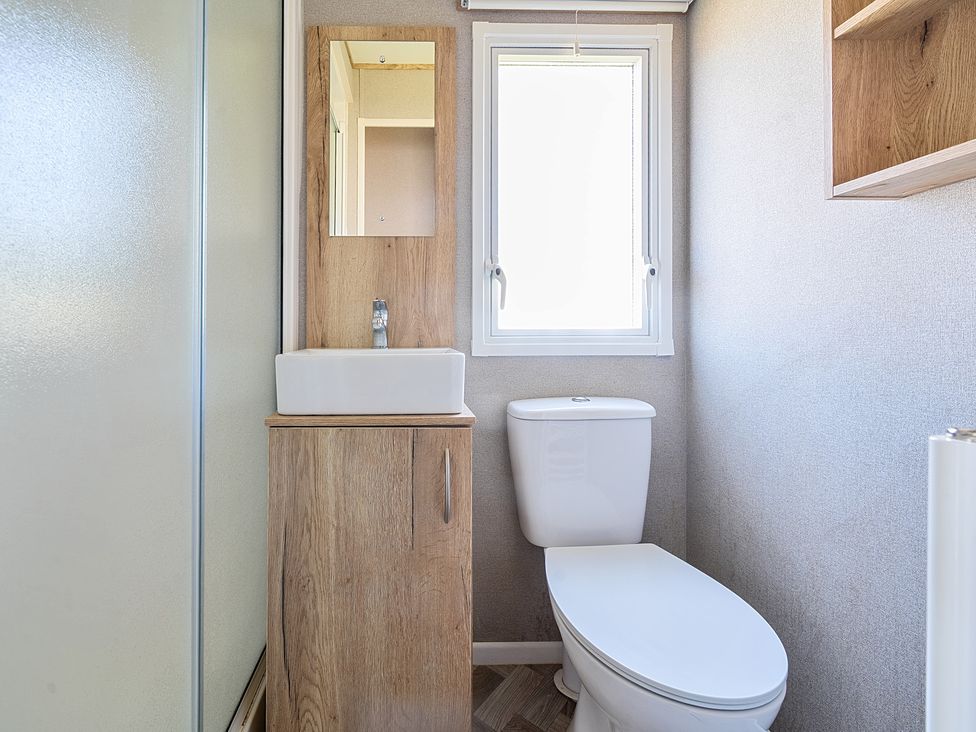 A bathroom with a toilet a sink with wooden cabinet a mirror and a window at Sunset Sands - Hayling Island in Hayling Island Holiday Park
