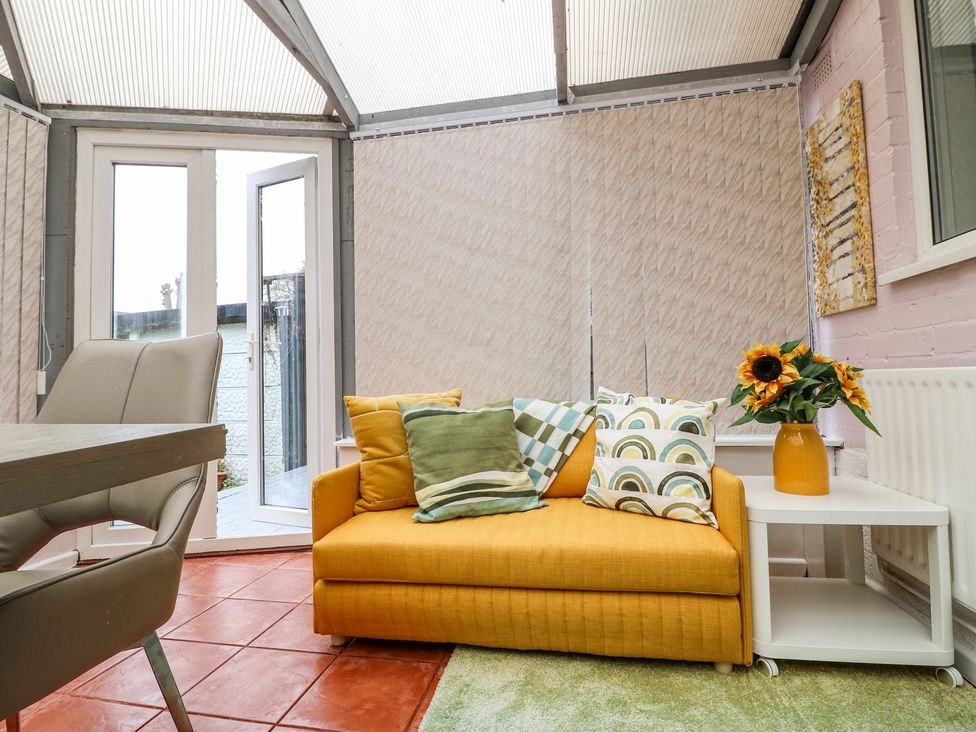 A conservatory with a yellow sofa with pillows and a white side table with sunflowers at 46 Dalby Road in Antsey