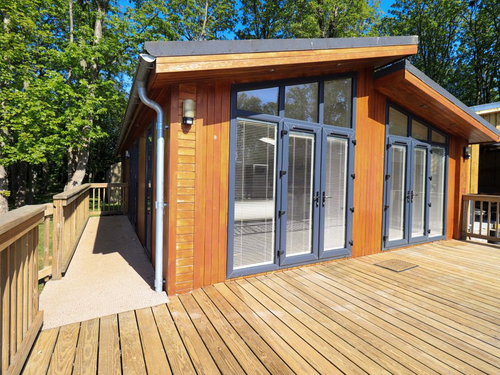 A wooden cabin with large glass doors and windows on a wooden deck surrounded by trees at Kingfisher Lodge in Gargrave