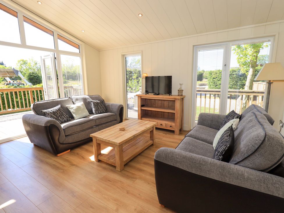 A living room with two gray sofas a wooden coffee table a wooden TV stand and large windows at Kingfisher Lodge in Gargrave