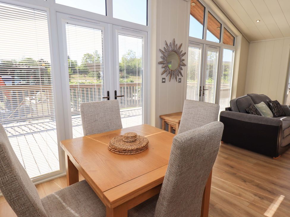 A dining table with four chairs and a round woven centerpiece next to large windows and a gray sofa in a living room at Kingfisher Lodge in Gargrave