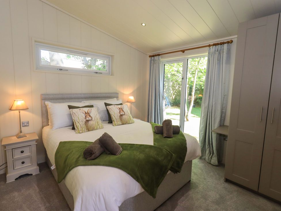 A bedroom with a double bed with green throws and rabbit cushions two lamps a wardrobe and sliding glass doors to the garden at Kingfisher Lodge in Gargrave