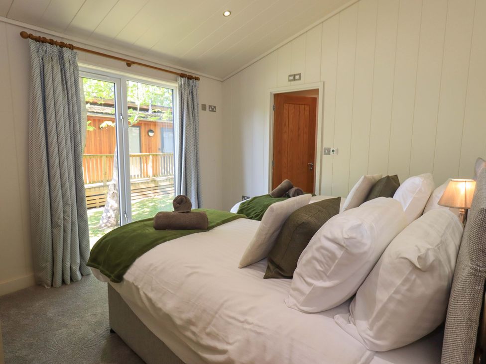A bedroom with two beds, pillows, rolled towels, a lamp, curtains over a glass door, and a wooden door at Kingfisher Lodge in Gargrave