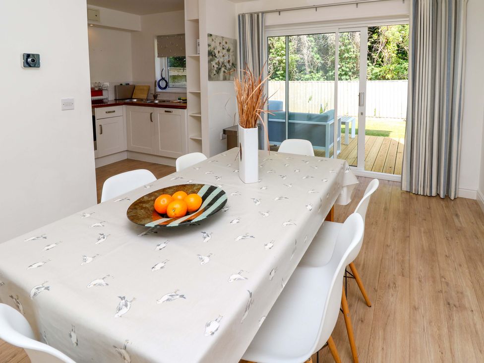 A dining area with a table covered by a cloth with bird patterns surrounded by white chairs and a bowl of oranges on the table with a kitchen and sliding glass doors leading to a patio outside at Whitfield P1 in Yarmouth
