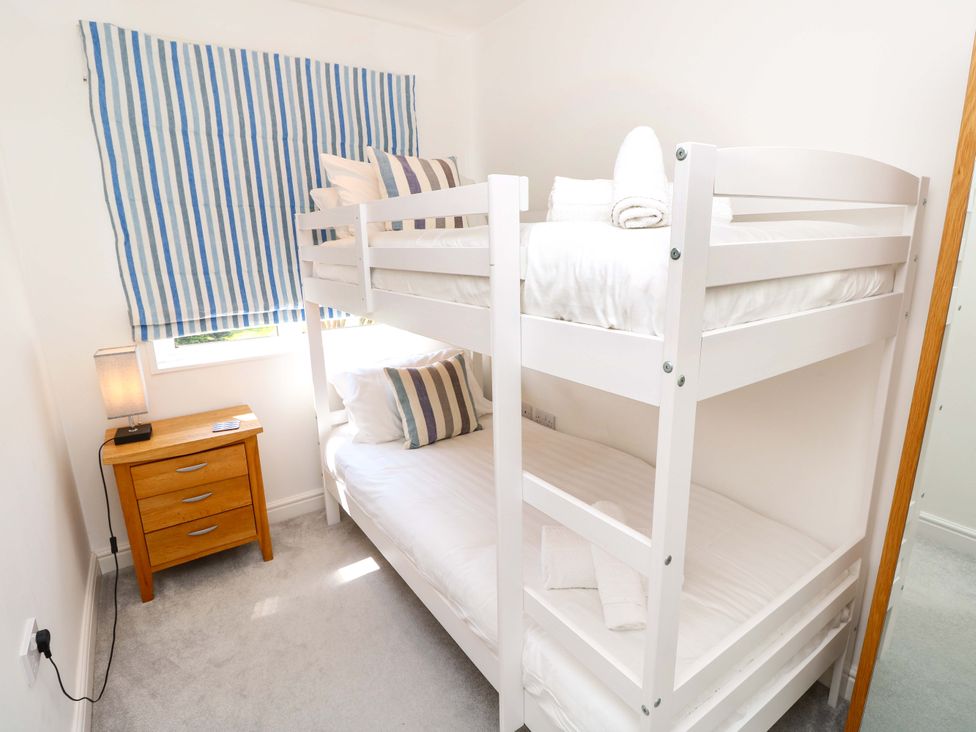 A bedroom with white bunk beds with striped pillows next to a wooden nightstand with a lamp and a window covered by a striped curtain at Whitfield P1 in Yarmouth