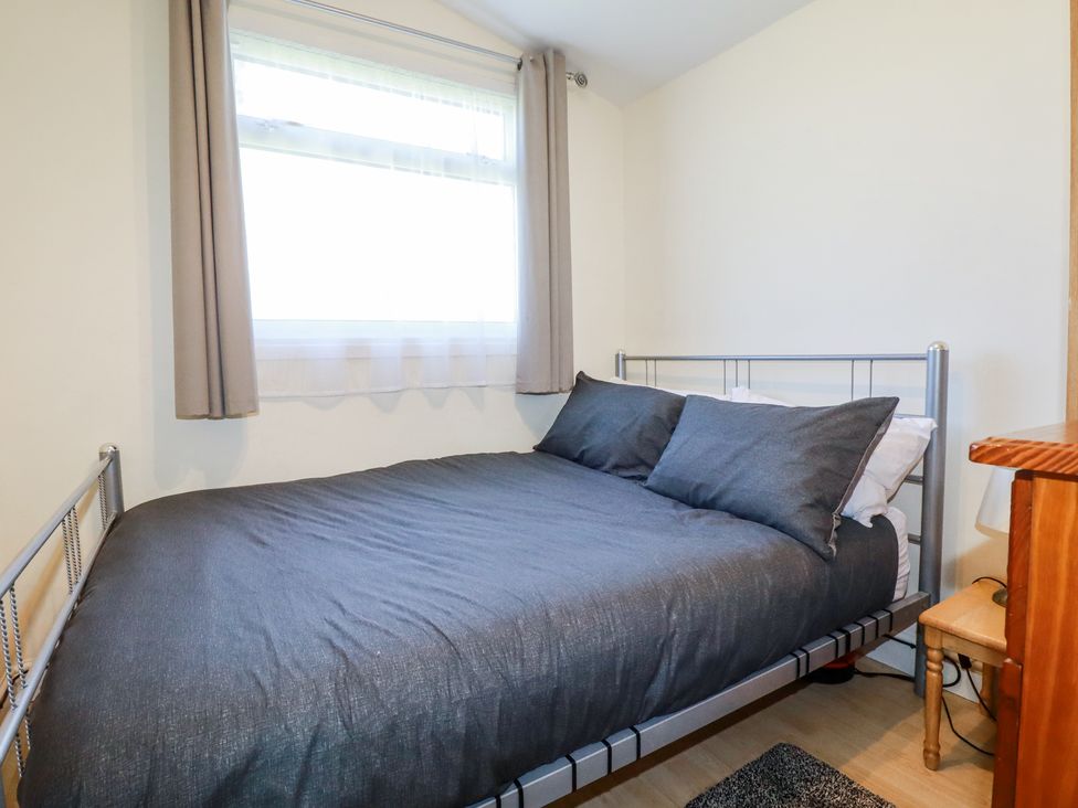 A bedroom with a double bed with dark grey bedding near a window with curtains and a wooden nightstand at 101 Atlantic Bays Holiday Park in Padstow