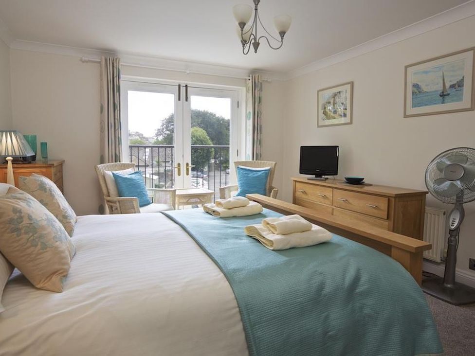 A bedroom with a bed covered in white and blue linens two chairs with blue cushions a wooden dresser with a television a standing fan and a lamp at 10 Dartmouth House in Dartmouth