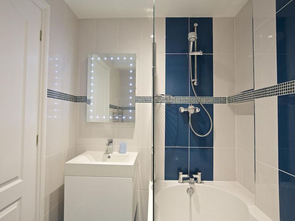 A bathroom with a white sink, a mirror with lights, a bathtub, and a shower with blue and white tiles at 10 Dartmouth House in Dartmouth