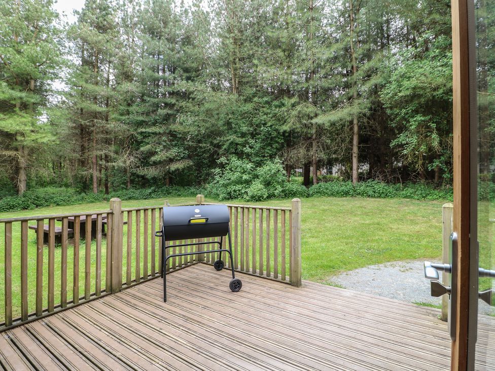 A wooden deck with a barbecue grill overlooking a grassy yard and forest at Sycamore Lodge in Rosliston