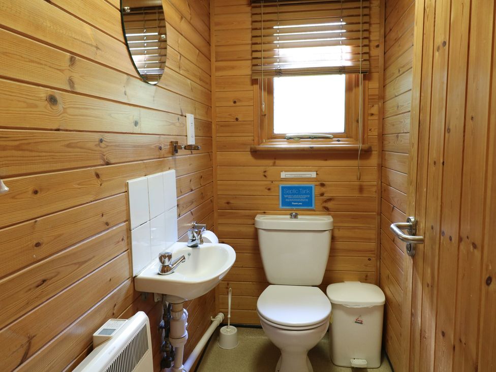 A small bathroom with a toilet, sink, mirror, window, trash bin, and wooden walls at Cedar Lodge in Rosliston