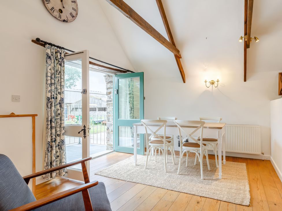 A dining area with a white table and four chairs on a rug near an open door with floral curtains and wooden beams on the ceiling at The Loft in Bodrean near Truro
