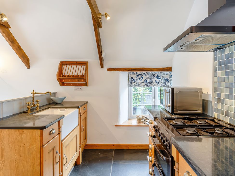 A kitchen with wooden cabinets a gas stove a microwave a farmhouse sink and a window with floral curtain at The Loft in Bodrean near Truro