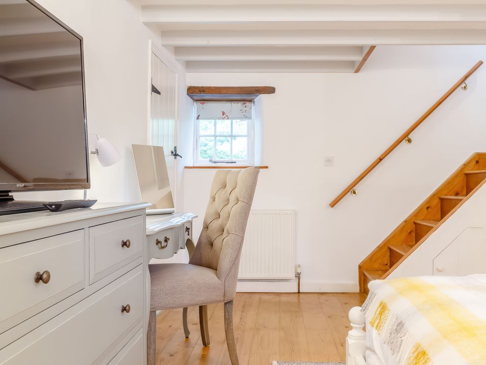 A bedroom with a white dresser holding a television a beige upholstered chair a wooden staircase and a small window at The Loft in Bodrean near Truro