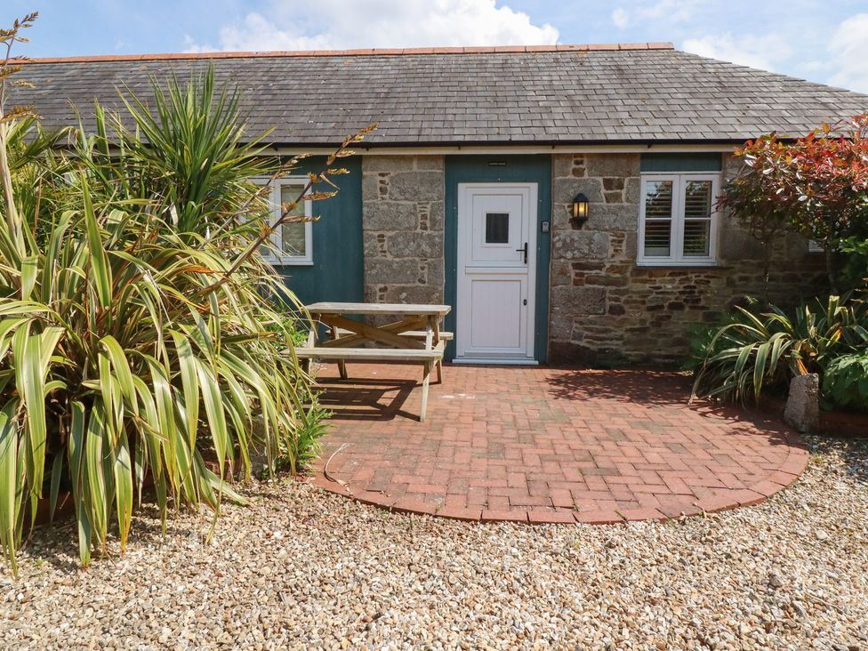 A stone building with a white door and windows a wooden picnic table on a brick patio and plants around the area at The Copper House in Bodrean near Truro