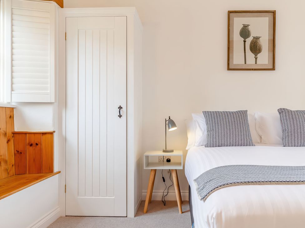 A bedroom corner with a white door wardrobe a bed with patterned pillows a small white bedside table with a lamp and wooden seat below a window at The Copper House in Bodrean near Truro