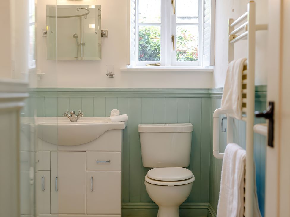 A bathroom with a white sink and cabinet, a toilet, a window with shutters, and a towel rack with towels at The Copper House in Bodrean near Truro