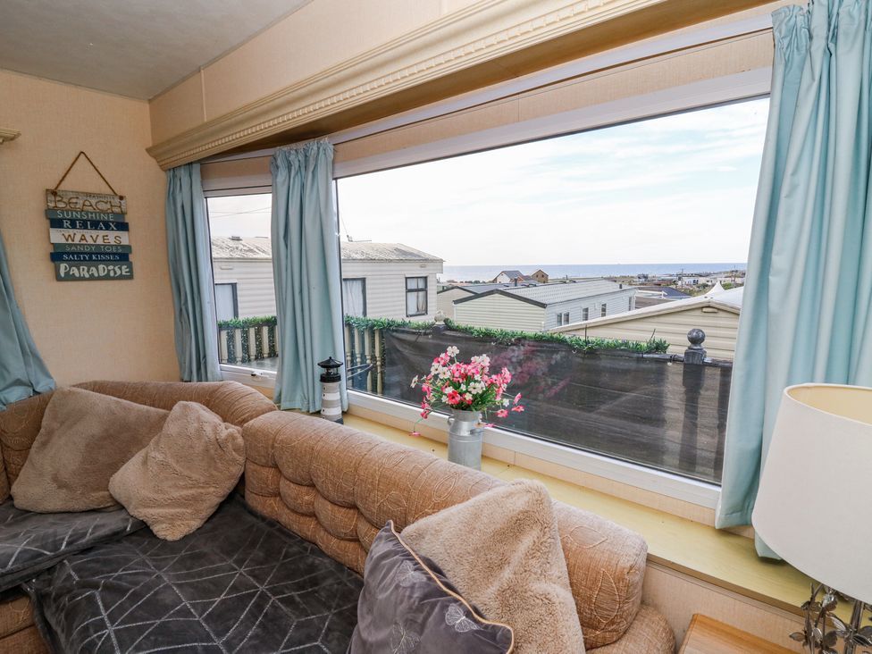A living room with a brown couch and pillows next to a large window with blue curtains overlooking other buildings at GM18 Clarach Bay near Aberystwyth