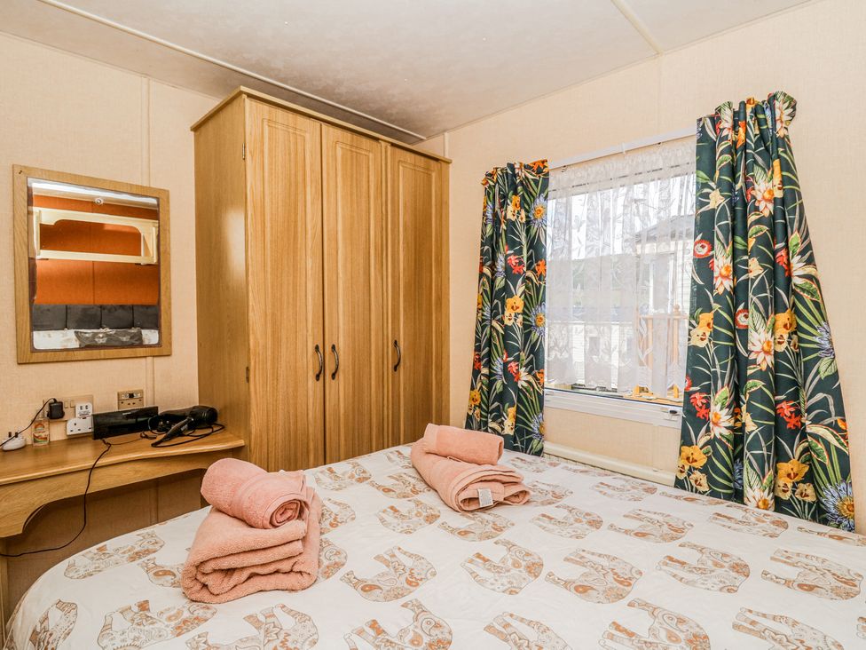 A bedroom with a bed covered with patterned bedspread and folded towels wardrobe window with floral curtains and a desk with mirror and hairdryer at GM18 Clarach Bay near Aberystwyth