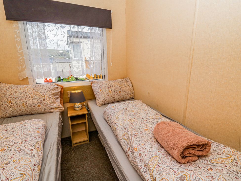 A small bedroom with two single beds a bedside table with a lamp and a window with lace curtains at GM18 Clarach Bay near Aberystwyth