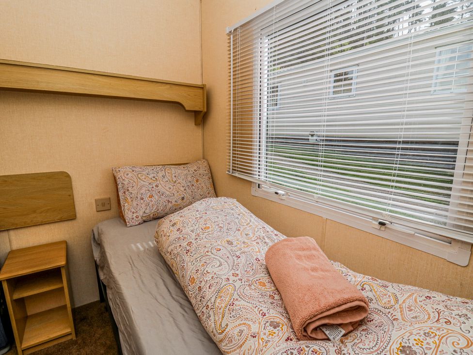A small bedroom with a single bed a patterned duvet and pillow a folded towel a wooden side table and blinds on the window at GM18 Clarach Bay near Aberystwyth