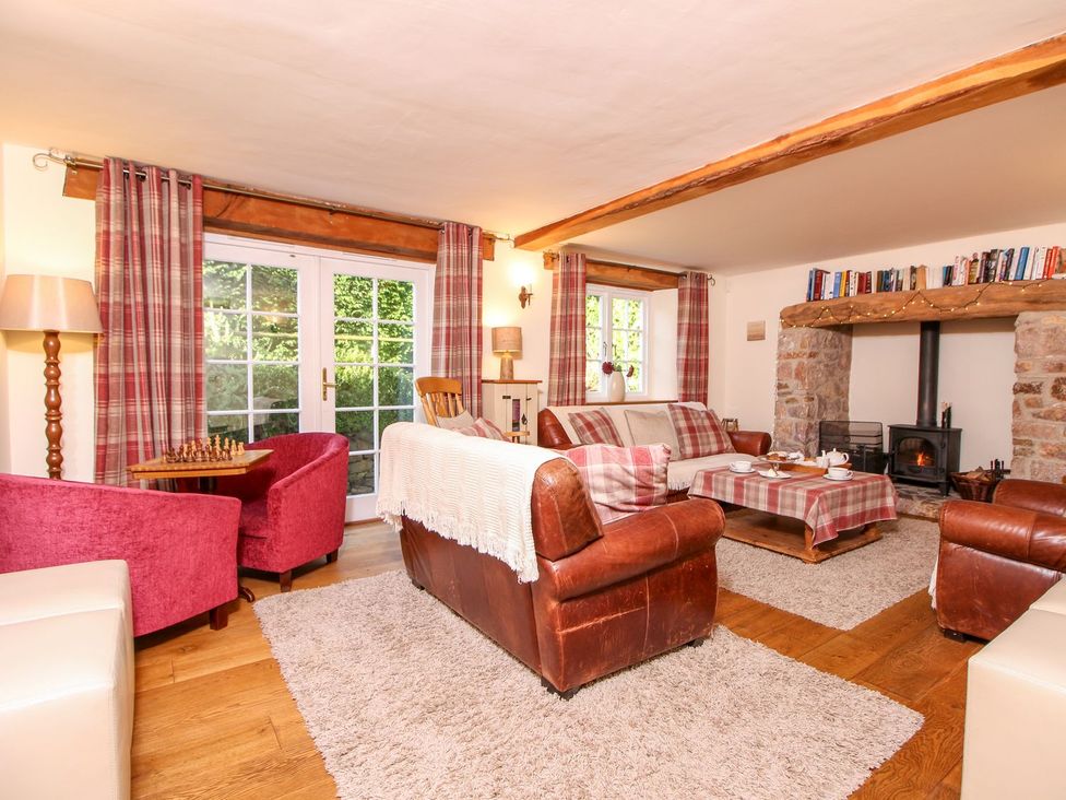 A living room with leather sofas, armchairs, a coffee table with a tablecloth, a stone fireplace, books on a wooden beam, and plaid curtains at Church Thatch in Abbotskerswell
