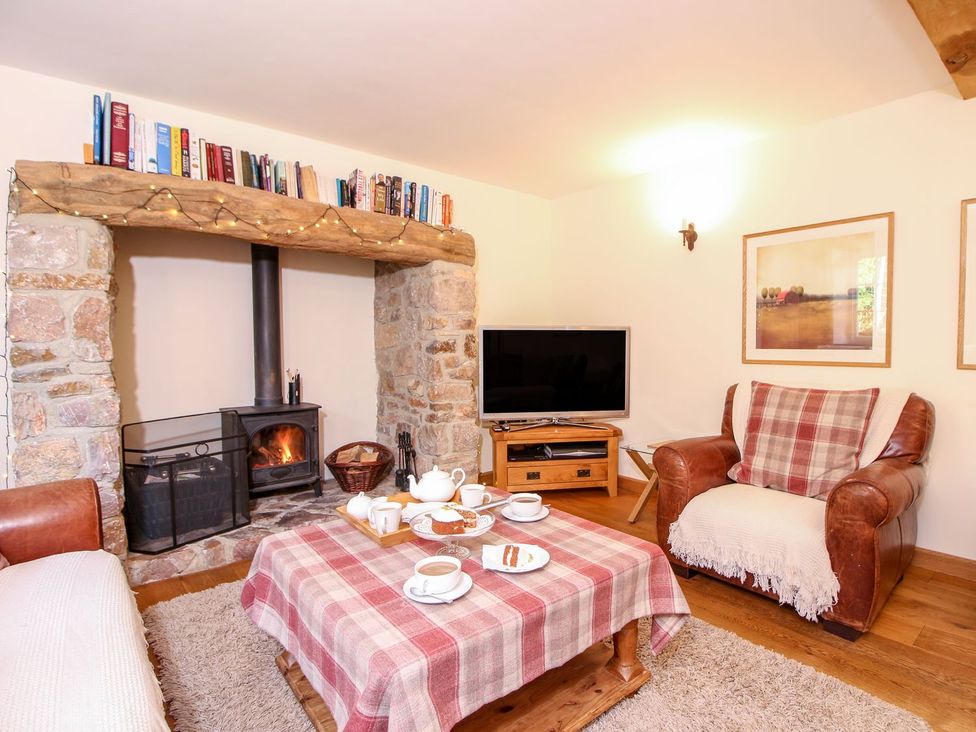 A living room with a stone fireplace a wood burning stove a television and a coffee table with tea and cake at Church Thatch in Abbotskerswell