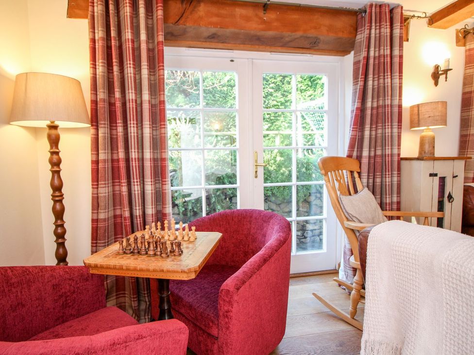 A seating area with two red armchairs and a wooden chess table by glass doors with red plaid curtains at Church Thatch in Abbotskerswell