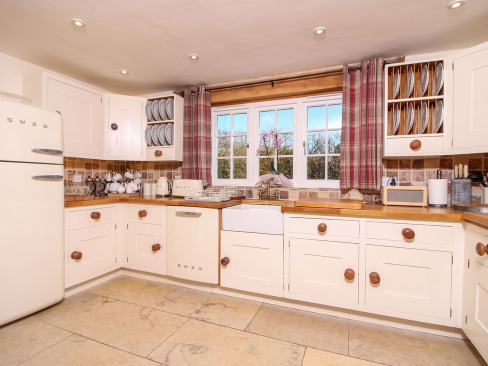 A kitchen with cream cabinets wooden countertops a farmhouse sink and a window with red plaid curtains at Church Thatch in Abbotskerswell