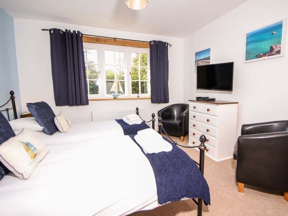 A bedroom with two single beds a window with dark curtains a chest of drawers and a wall mounted television at Church Thatch in Abbotskerswell