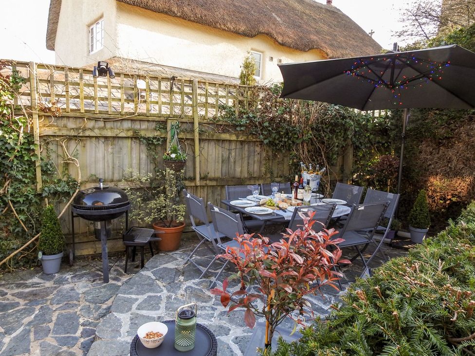 An outdoor patio with a dining table set with plates and drinks surrounded by chairs a barbecue grill and a large umbrella with string lights at Church Thatch in Abbotskerswell