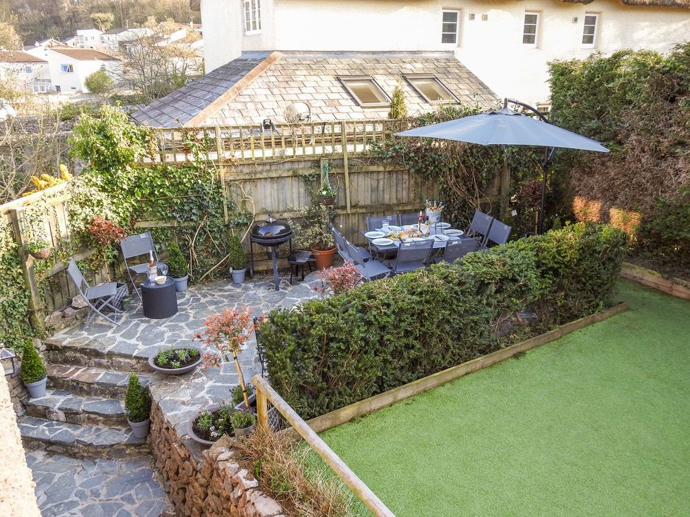 A garden patio with a dining table set under an umbrella surrounded by chairs and a barbecue grill at Church Thatch in Abbotskerswell