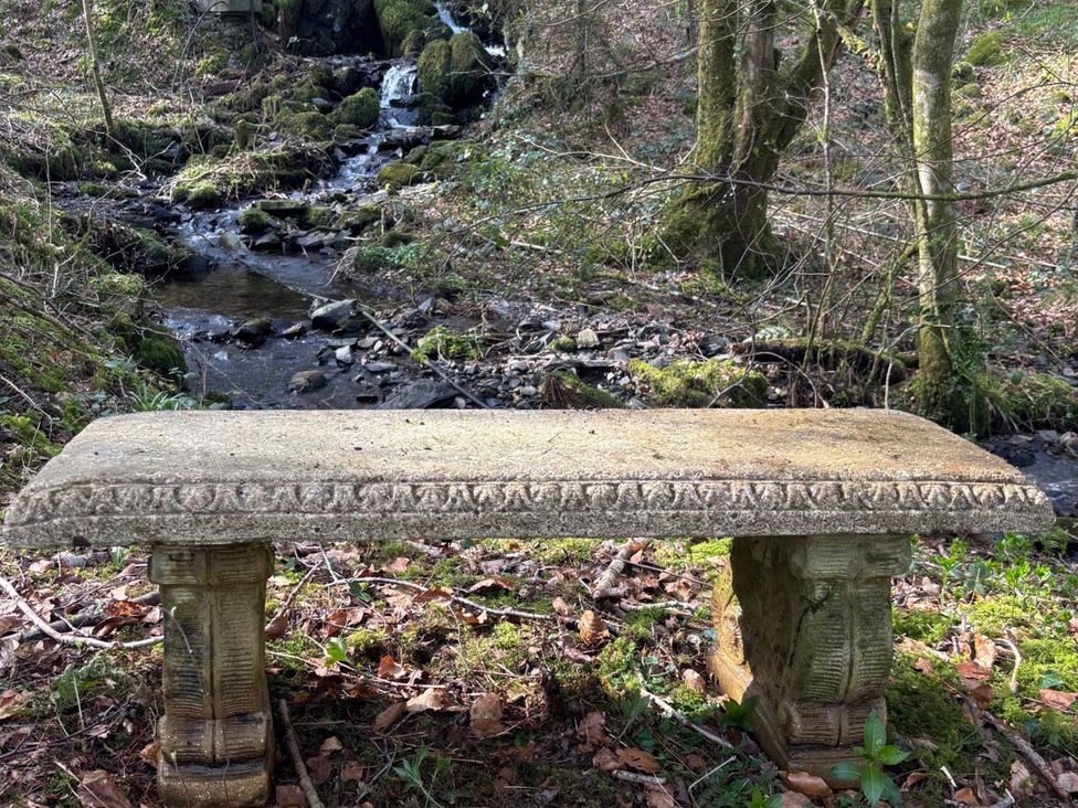 A bench near a stream with trees in the background at Bracken Ground Coniston