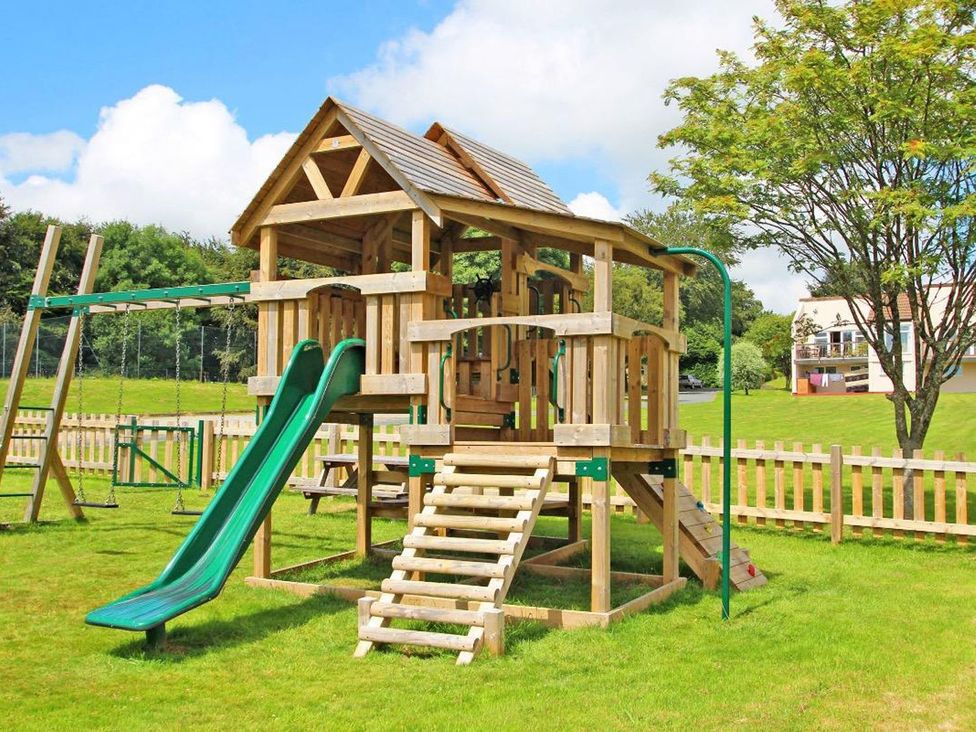 A playhouse with a slide and swings in an outdoor play area at Valley Lodge 2 Callington