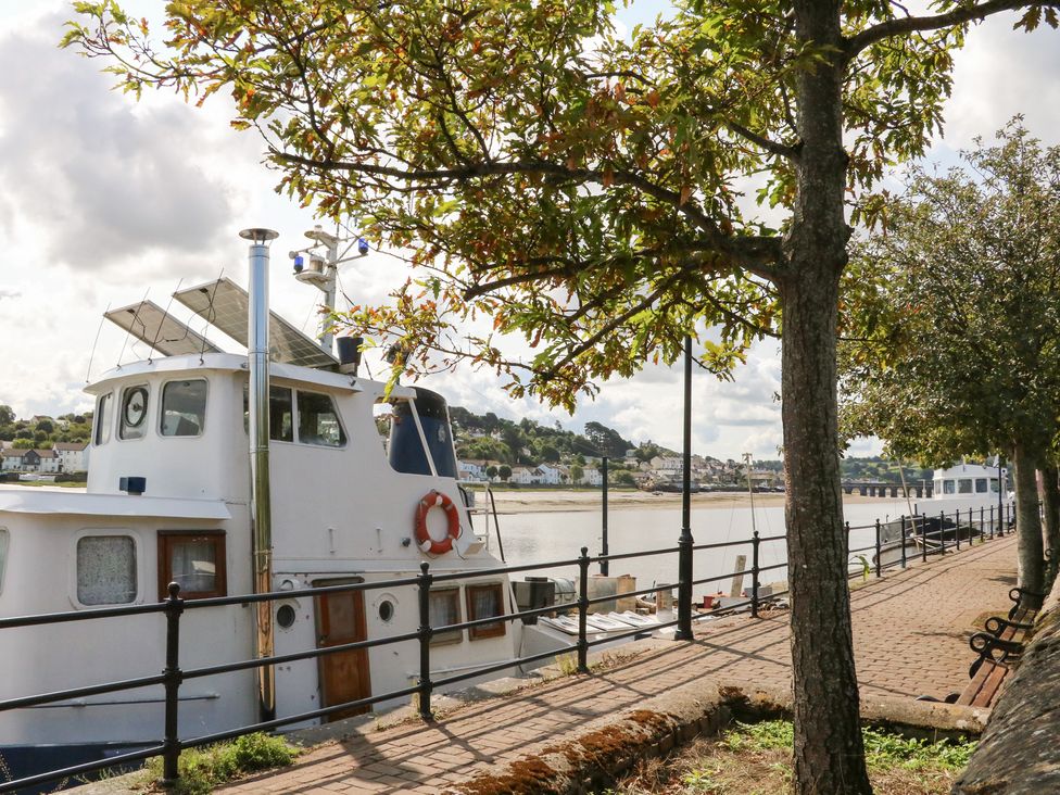 A boat docked at a river with trees and a pathway at Quay Close Cottage