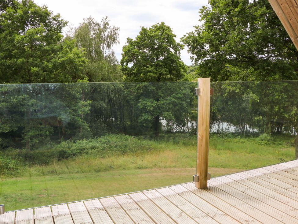 A wooden deck with a glass railing overlooking a grassy area and trees at Lakeside Lodge near Burwash