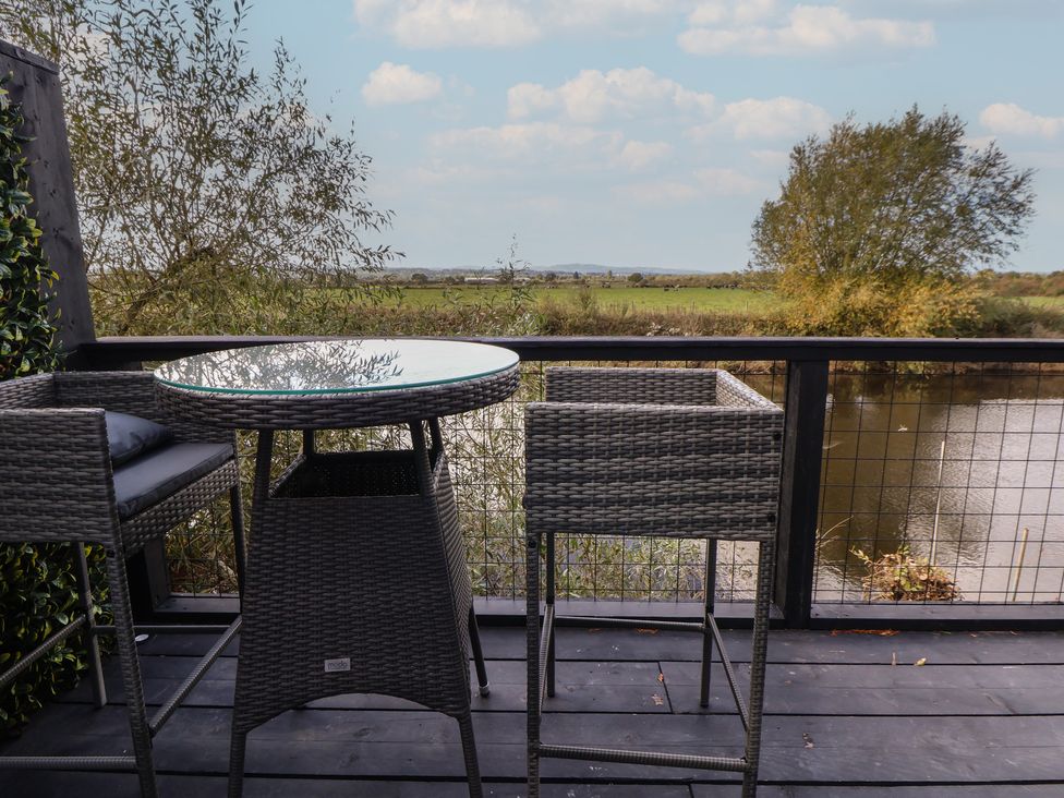 A table and stools on a balcony overlooking water and grass at The Larch House in Farndon