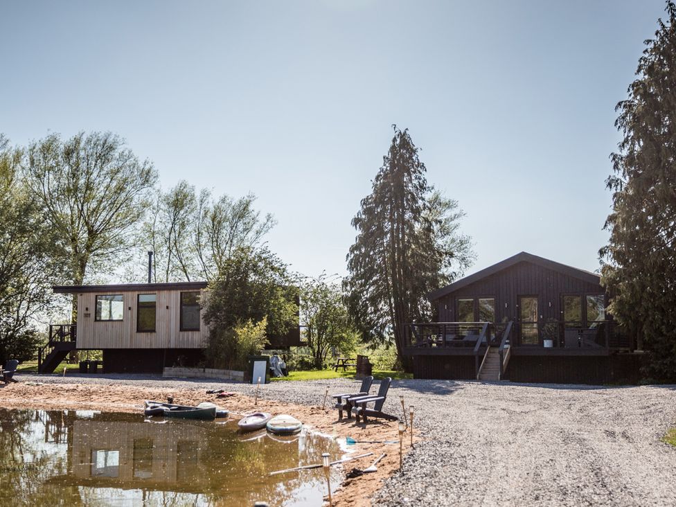 Two houses by the water with trees and gravel area at The Larch House in Farndon