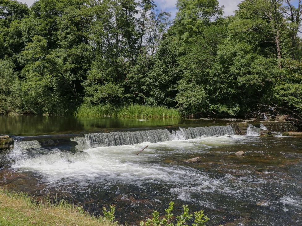 A river with a waterfall flowing over rocks and surrounded by trees and grass at Railway Retreat at Eamont Park in Penrith