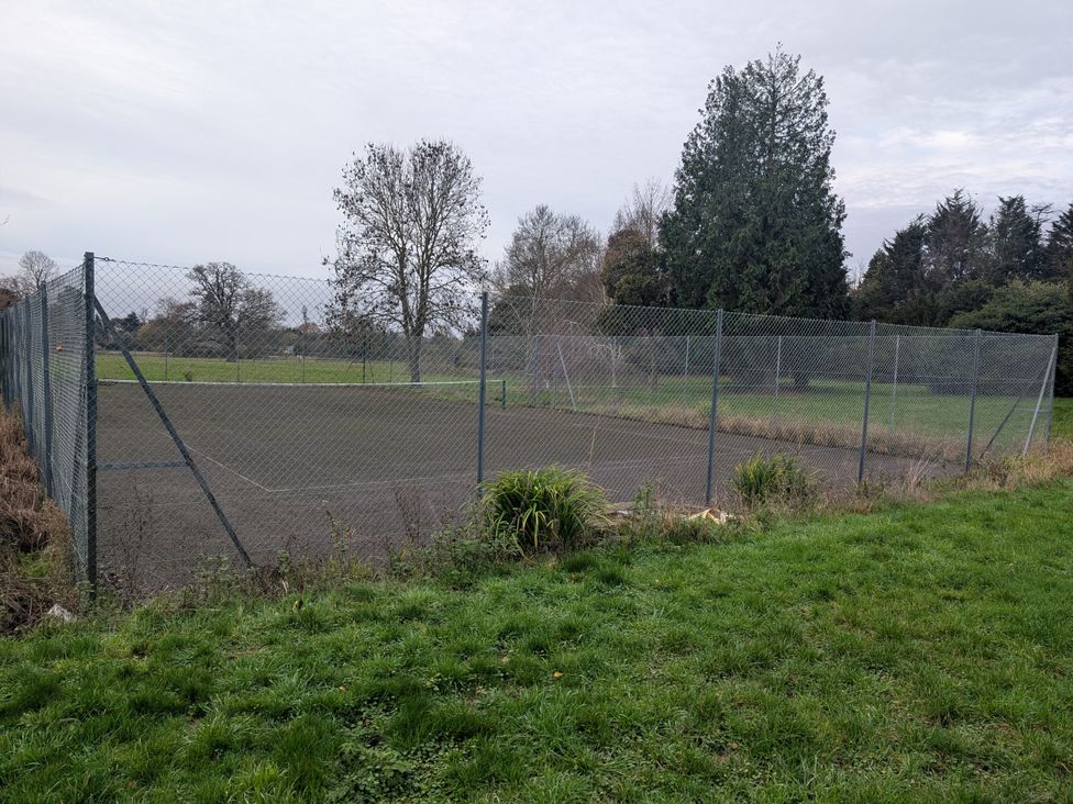 A tennis court enclosed by a fence with grass and trees at Heath Lodge in Iver