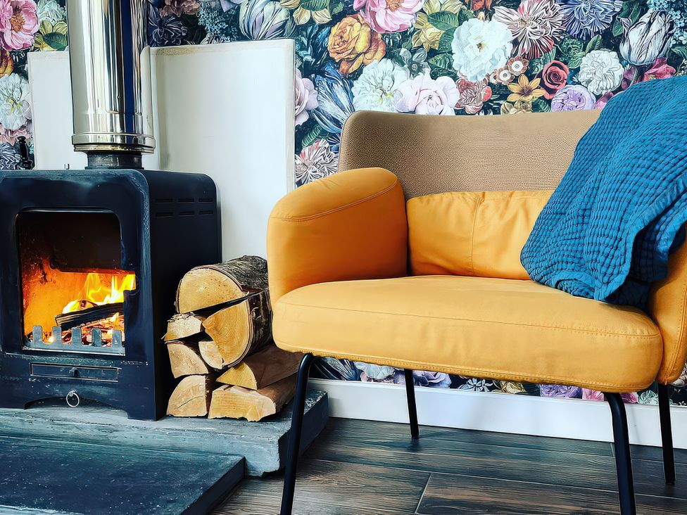 A living room with a stove, chair, and firewood at Hen Stablau Dinmael near Cerrigydrudrion