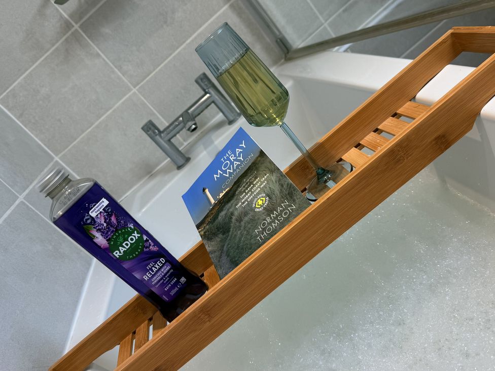 A bathtub with a bamboo tray holding a drink and a book at Portknockie Cottage in Buckie