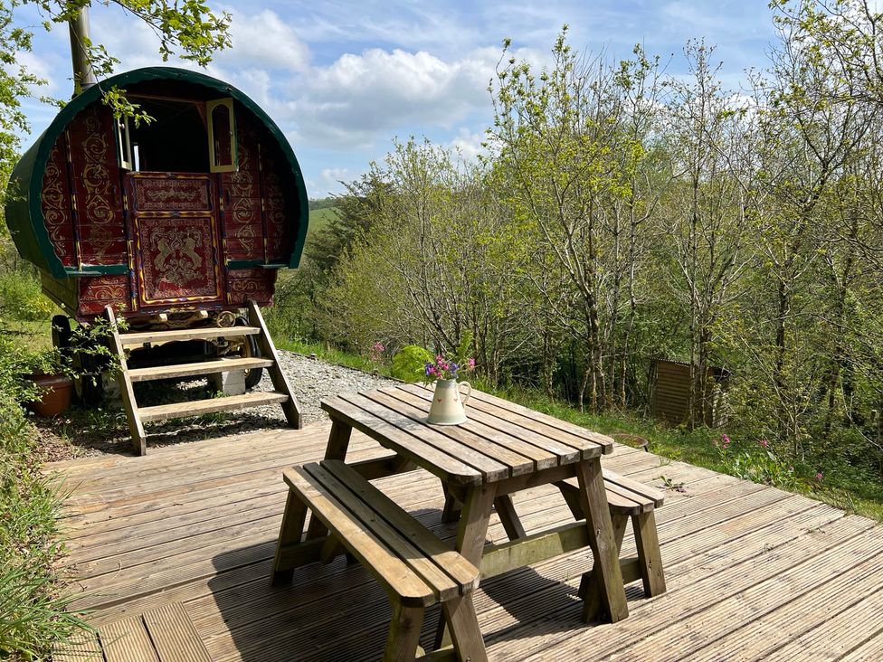 An outdoor seating area with a wooden table and benches beside a gypsy caravan at Goldfinch in Panson Mill near Ashwater