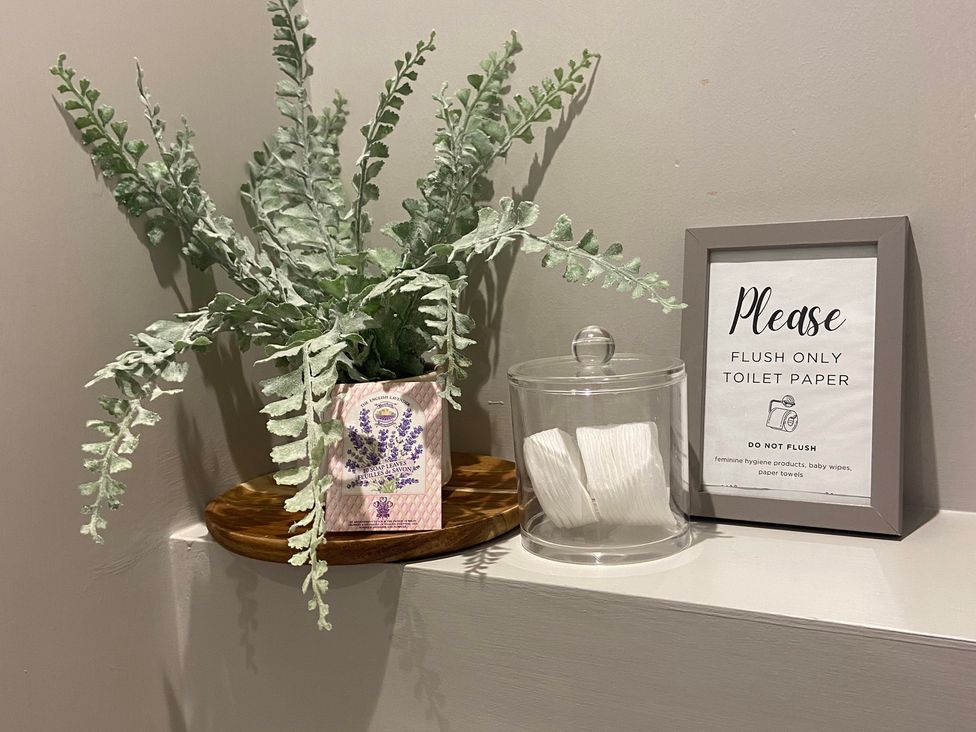 A bathroom display with a fake plant, decorative box, glass jar with tissue paper and a warning sign at The Cart Shed in Bedford