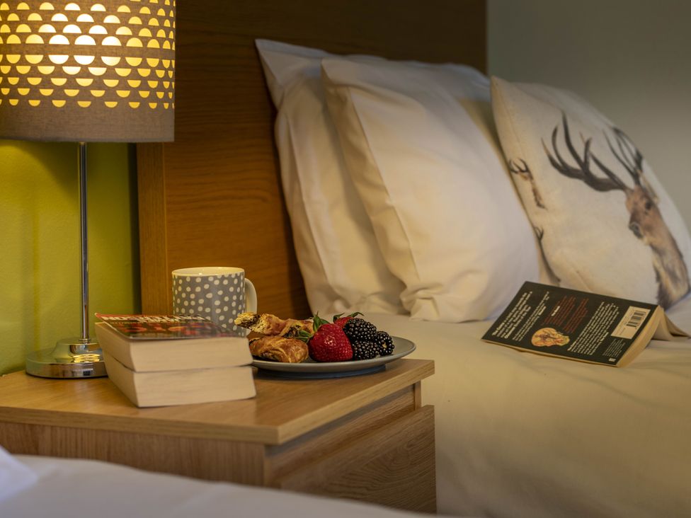A bedroom with a bedside table featuring a lamp, books, a cup, and a plate of food at Clayhills Cottage in Blairgowrie