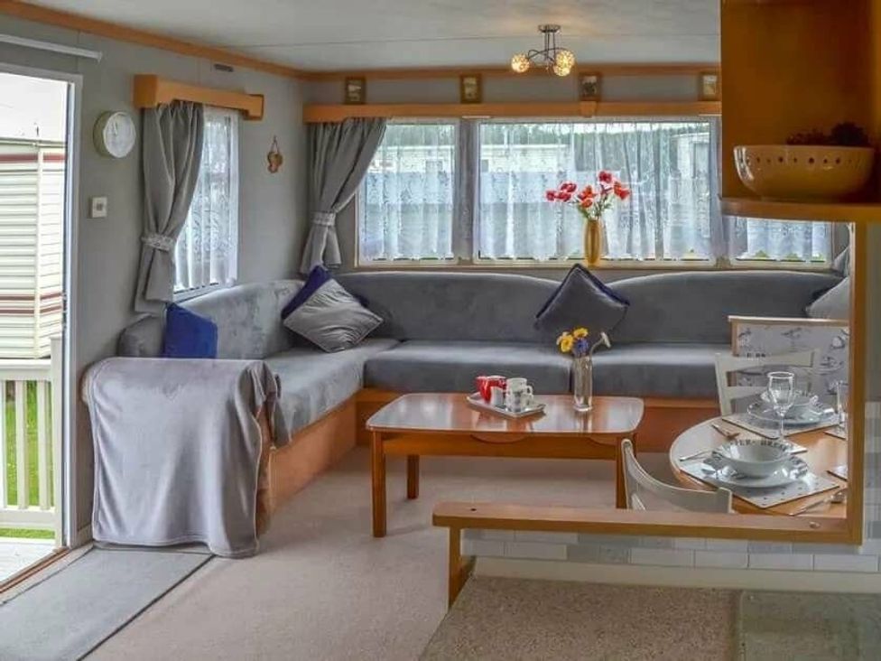 a living room with a grey sofa, a wooden table with dishes, and curtains at Beach Haven Caravan Park - Holiday Accommodation 16700, Bacton