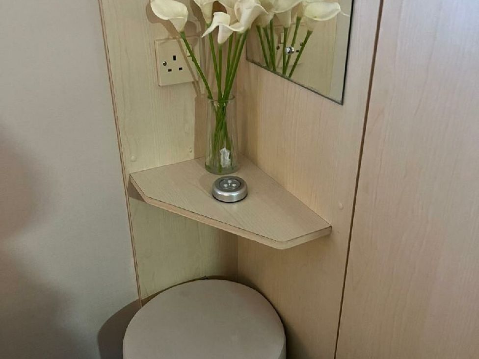 a corner shelf with a vase of flowers and a small mirror above a round stool at Three Lochs Holiday Park - Holiday Accommodation 17424 in Stranraer