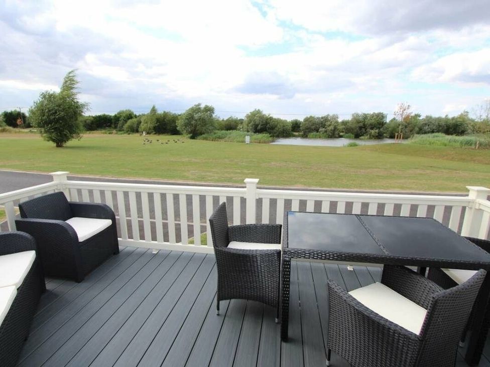 a patio area with black wicker chairs and a table overlooking a grassy field and pond at Tattershall Lakes Country Park - Holiday Accommodation 17807 in Tattershall